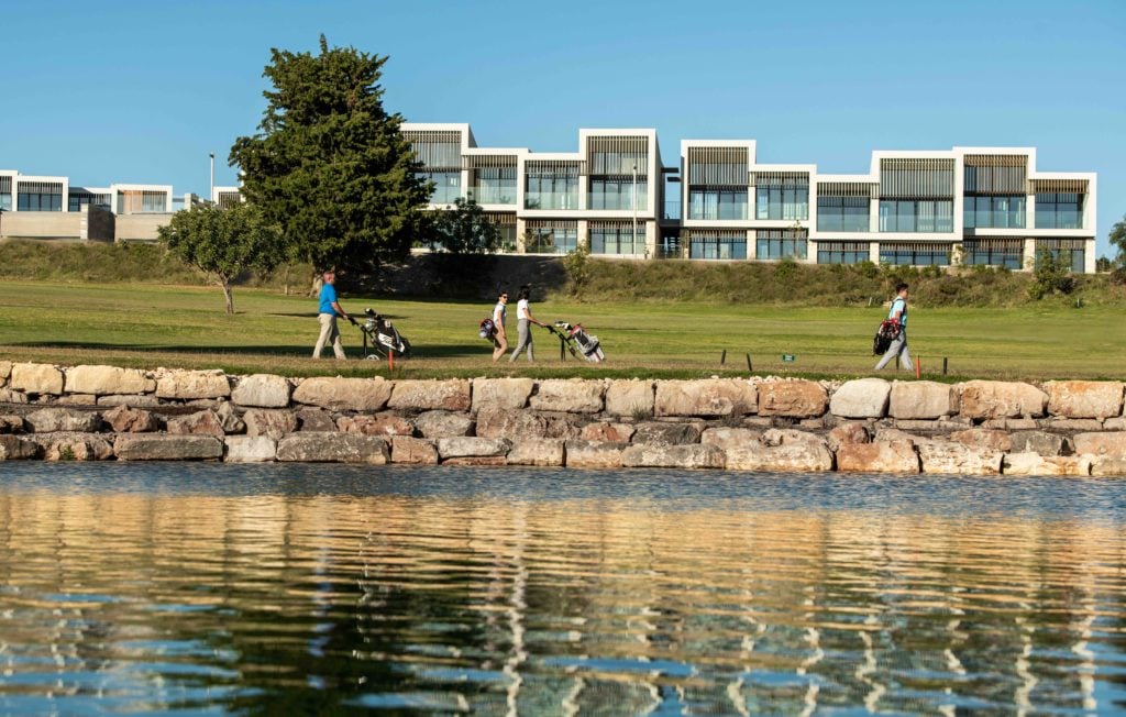Golf-_-Suites-1024x652jpeg - Sovereign Estates Sovereign Estates Golfers walk along a grassy lakeside path with their golf bags stone seawall in the foreground and modern buildings in the background 2026 Golfers walk along a grassy lakeside path with their golf bags stone seawall in the foreground and modern buildings in the background