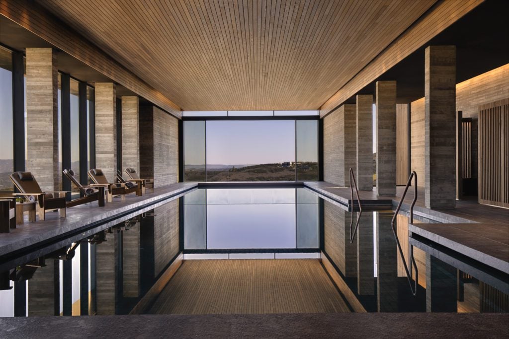 Minthis-Spa-Pool-View-1024x683jpg - Sovereign Estates Sovereign Estates Indoor resort pool with stone columns wooden ceiling and row of lounge chairs facing a desert view through large windows 2026 Indoor resort pool with stone columns wooden ceiling and row of lounge chairs facing a desert view through large windows