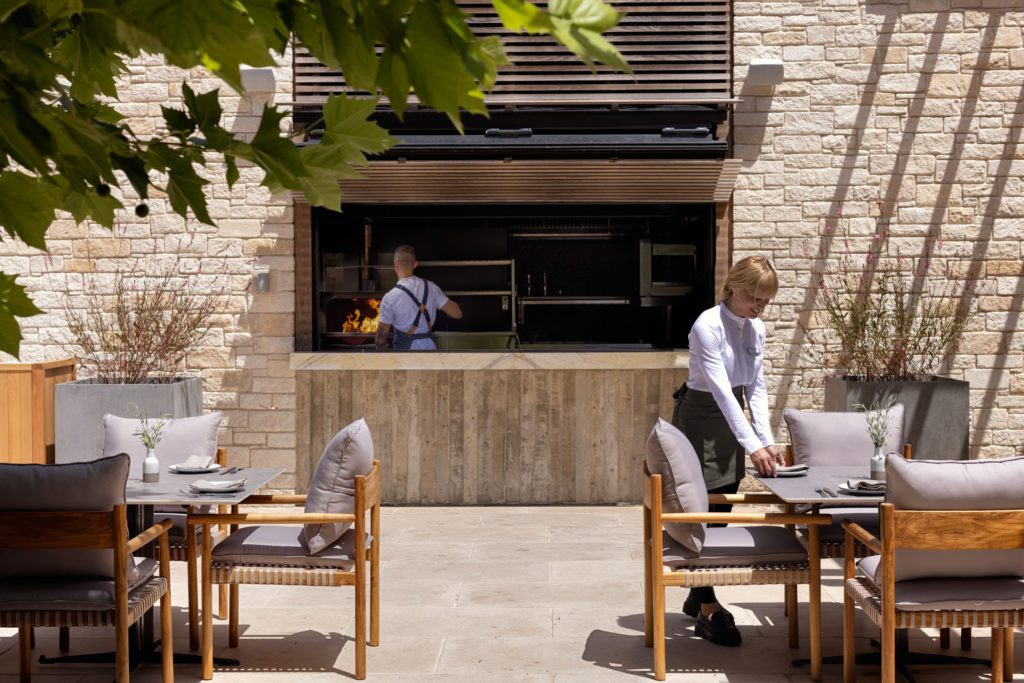 Restaurant-1024x683jpg - Sovereign Estates Sovereign Estates Outdoor restaurant patio with wooden tables and cushioned chairs a server sets a table while a chef grills at an open kitchen in the background 2026 Outdoor restaurant patio with wooden tables and cushioned chairs a server sets a table while a chef grills at an open kitchen in the background
