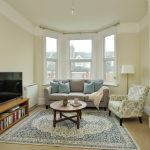 Sovereign Estates   Cozy living room with beige walls bay windows a gray sofa with blue cushions a patterned armchair a round wooden coffee table TV on a stand and a blue patterned rug 2026 Cozy living room with beige walls bay windows a gray sofa with blue cushions a patterned armchair a round wooden coffee table TV on a stand and a blue patterned rug