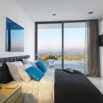 Sovereign Estates   Modern hotel bedroom with a king bed blue accent pillows and a wall mounted TV overlooking a scenic valley through floor to ceiling glass doors 2026 Modern hotel bedroom with a king bed blue accent pillows and a wall mounted TV overlooking a scenic valley through floor to ceiling glass doors