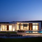 Sovereign Estates   Modern single story home at dusk with stone facade large glass walls and illuminated interior beside a rectangular pool 2026 Modern single story home at dusk with stone facade large glass walls and illuminated interior beside a rectangular pool