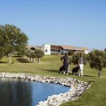 Sovereign Estates   Two men in casual clothes walk across a sunny golf course each with a golf bag near a rocky pond and a modern clubhouse in the background 2026 Two men in casual clothes walk across a sunny golf course each with a golf bag near a rocky pond and a modern clubhouse in the background