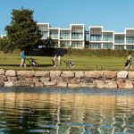 Sovereign Estates   People with golf bags walking along a grassy park beside a rocky seawall modern white buildings in the background and calm water in the foreground 2026 People with golf bags walking along a grassy park beside a rocky seawall modern white buildings in the background and calm water in the foreground