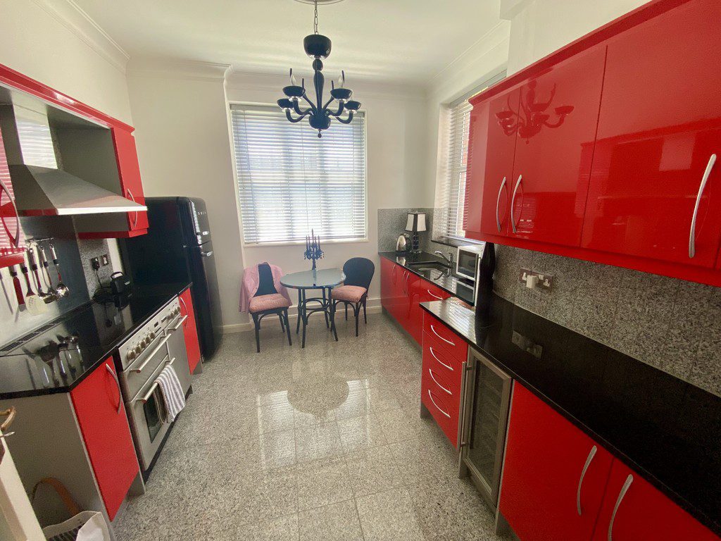 IMG_21_3_largejpg - Sovereign Estates Sovereign Estates Modern kitchen with glossy red cabinets black countertops and stainless steel appliances a small round dining table and two chairs sit by a window with blinds under a black chandelier 2026 Modern kitchen with glossy red cabinets black countertops and stainless steel appliances a small round dining table and two chairs sit by a window with blinds under a black chandelier