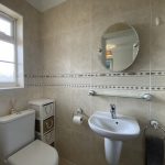 Sovereign Estates   Bright beige tiled bathroom with a white toilet pedestal sink circular mirror glass shelf and a small chest of drawers beside the toilet 2026 Bright beige tiled bathroom with a white toilet pedestal sink circular mirror glass shelf and a small chest of drawers beside the toilet