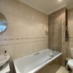 Sovereign Estates   Modern beige tiled bathroom with a white bathtub and clear glass shower screen a wall mirror sink and toilet plus open shelving with towels on the right side 2026 Modern beige tiled bathroom with a white bathtub and clear glass shower screen a wall mirror sink and toilet plus open shelving with towels on the right side