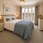 Sovereign Estates   Bedroom with a white quilted bed blue throw and pillows two bedside lamps and glass doors to a balcony with curtains 2026 Bedroom with a white quilted bed blue throw and pillows two bedside lamps and glass doors to a balcony with curtains