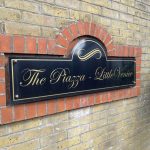IMG_23_19_largejpg - Sovereign Estates Sovereign Estates Black rectangular sign mounted on a brick wall reads The Piazza Little Venice in gold script with decorative arch above 2026 Black rectangular sign mounted on a brick wall reads The Piazza Little Venice in gold script with decorative arch above