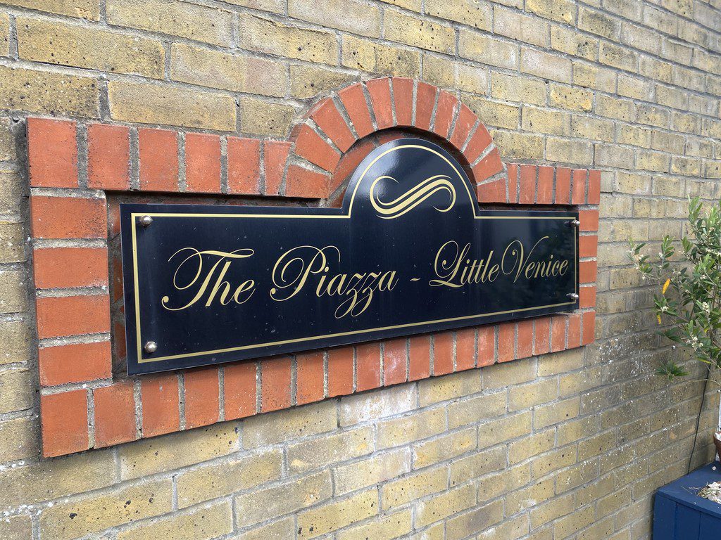 IMG_23_19_largejpg - Sovereign Estates Sovereign Estates Black rectangular sign mounted on a brick wall reads The Piazza Little Venice in gold script with decorative arch above 2026 Black rectangular sign mounted on a brick wall reads The Piazza Little Venice in gold script with decorative arch above