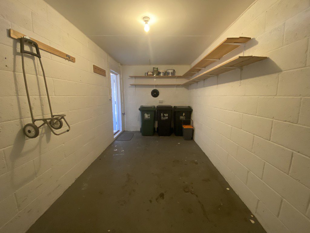 IMG_23_37_largejpg - Sovereign Estates Sovereign Estates Narrow garage with white cinder block walls a single ceiling light wall shelves on the right and twothree trash bins at the far end a hand truck hangs on the left wall 2026 Narrow garage with white cinder block walls a single ceiling light wall shelves on the right and twothree trash bins at the far end a hand truck hangs on the left wall