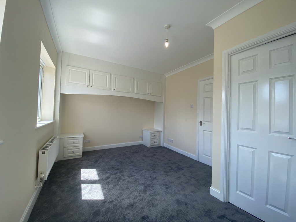 IMG_23_65_largejpg - Sovereign Estates Sovereign Estates Bright empty bedroom with built in white cupboards above a desk alcove beige walls and gray carpet window and radiator on the left 2026 Bright empty bedroom with built in white cupboards above a desk alcove beige walls and gray carpet window and radiator on the left