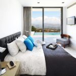 Sovereign Estates   Bright hotel room with a king bed white linens blue throw pillows and a gray tufted headboard facing glass doors to a pool and mountain view 2026 Bright hotel room with a king bed white linens blue throw pillows and a gray tufted headboard facing glass doors to a pool and mountain view