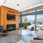 Sovereign Estates   Bright contemporary living room with a stone accent wall wood paneled fireplace surround mounted TV and a glass coffee table facing a beige sofa and large glass doors to a patio area 2026 Bright contemporary living room with a stone accent wall wood paneled fireplace surround mounted TV and a glass coffee table facing a beige sofa and large glass doors to a patio area