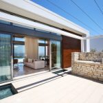 Sovereign Estates   Patio of a modern home with sliding glass doors to a living room beige sofa visible and a stone outdoor grilling area on the terrace with a distant ocean view 2026 Patio of a modern home with sliding glass doors to a living room beige sofa visible and a stone outdoor grilling area on the terrace with a distant ocean view
