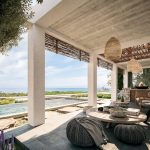 Sovereign Estates   Coastal outdoor living room with white columns woven pendant lights and a seaview lounge area under a shaded patio 2026 Coastal outdoor living room with white columns woven pendant lights and a seaview lounge area under a shaded patio