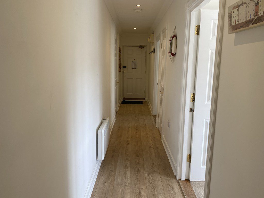 IMG_25_41_largejpg - Sovereign Estates Sovereign Estates Narrow white hallway with light wood floor leading to a closed front door a lifebuoy hangs on the right wall at mid height 2026 Narrow white hallway with light wood floor leading to a closed front door a lifebuoy hangs on the right wall at mid height